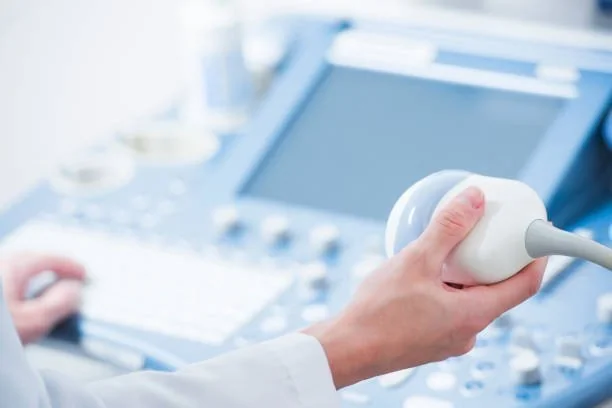 Mão de profissional de saúde segurando transdutor de ultrassom, com aparelho de ultrassonografia ao fundo